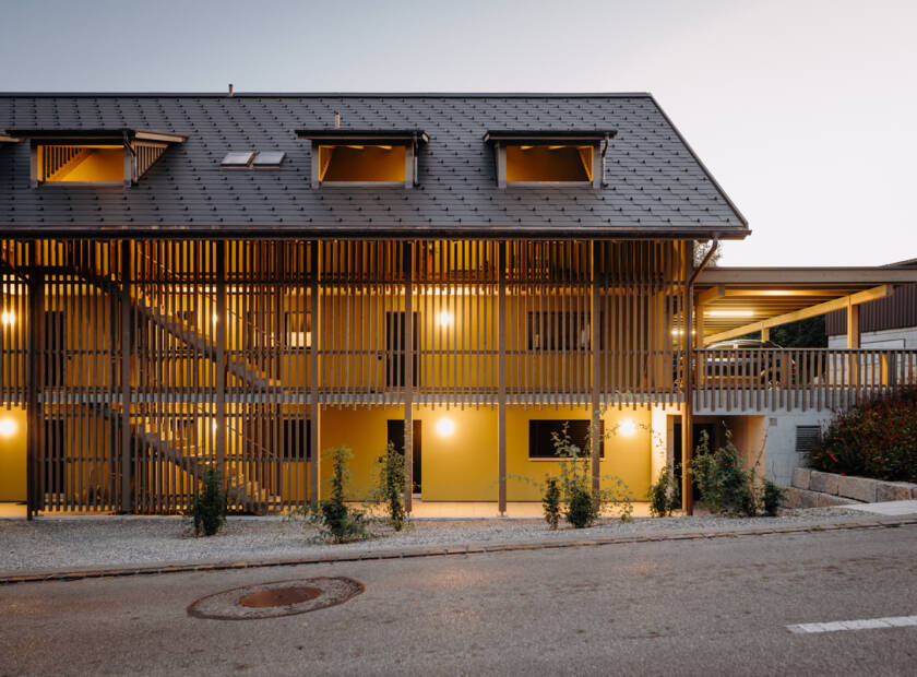 Mehrfamilienhaus in Hausform aus Holz und mit Solardach bei Tag und bei Dämmerung MFH, Hüttwilen, CH